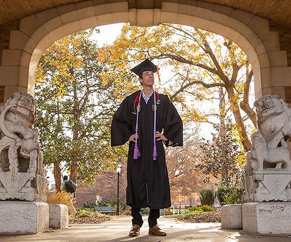 Student wearing cap and gown standing between lion statues under journalism archway